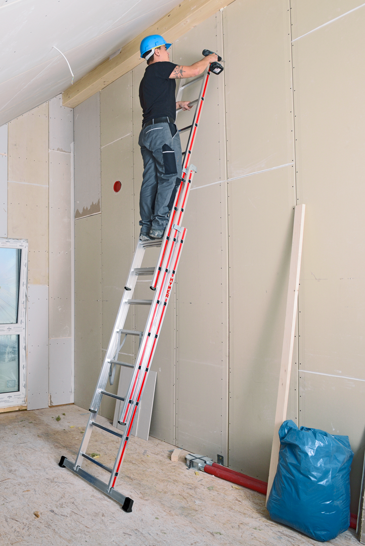Plaquiste installé sur une échelle transformable à la bonne hauteur pour travailler en sécurité, fixant des plaques de plâtres